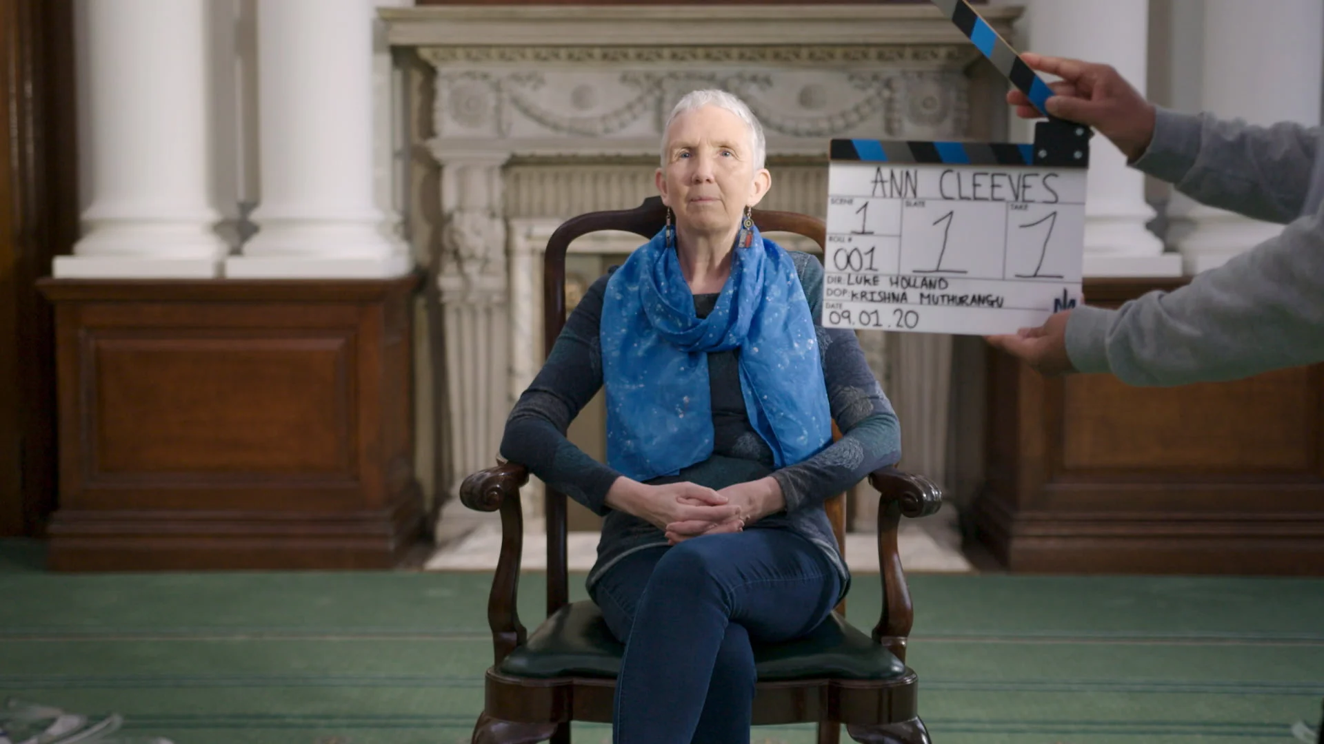 Ann Cleeves Sat, Centrally Framed In A Large Room. A Clapperboard Is Visible In The Right Of Frame.