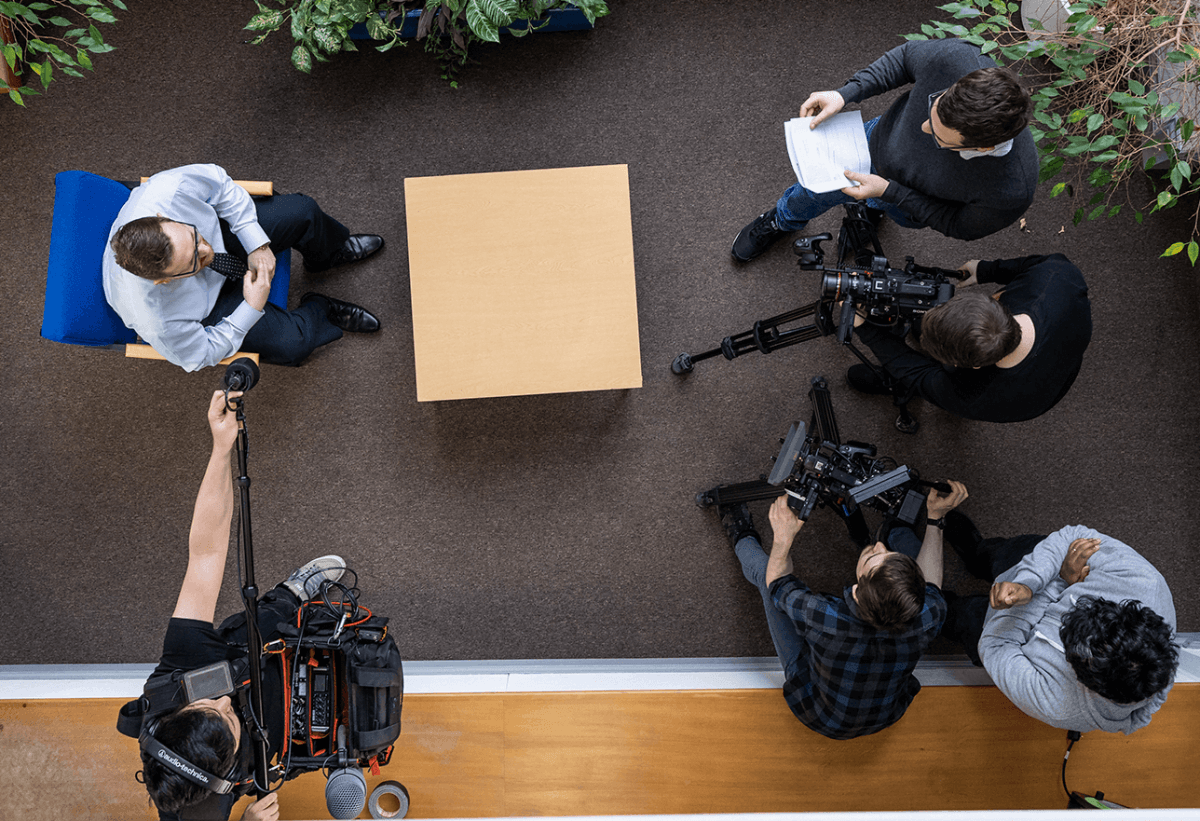 A corporate video production film crew as seen from overhead. Two camera operators, a director, a producer, a sound recordist and an interviewee can be seen in an office setting.