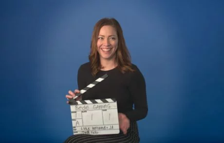A Woman Sat In Front Of A Blue Screen, Laughing Whilst Holding A Clapperboard