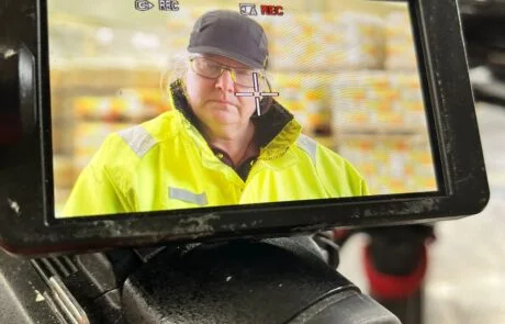 A Woman Featuring In A Manufacturing Video , Stood In Front Of A Large Number Of Pallets Stocked With Adhesive.