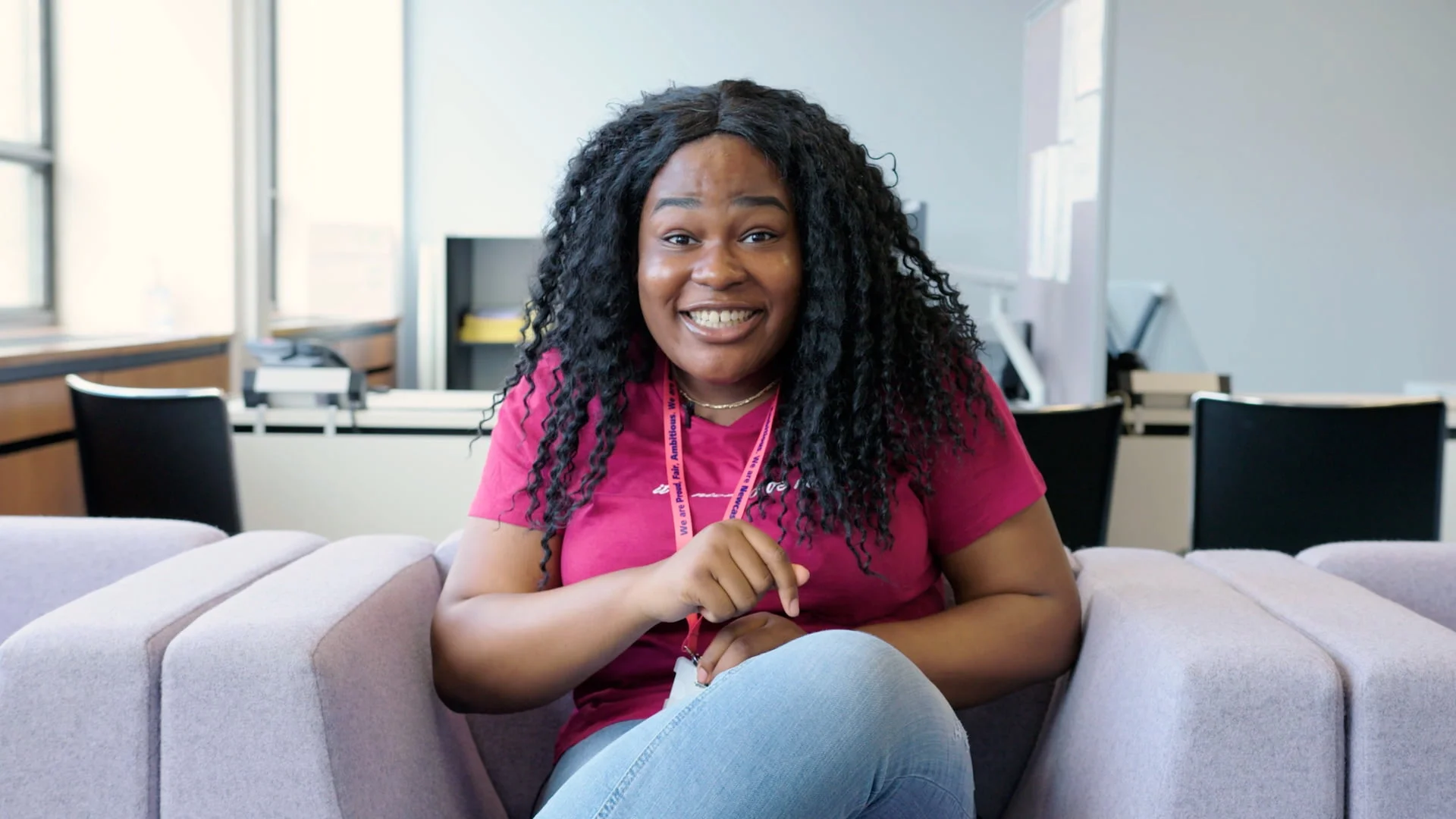 A Young Woman Is Sat In An Office, She Is Centrally Framed And Looking Directly Into The Lens.