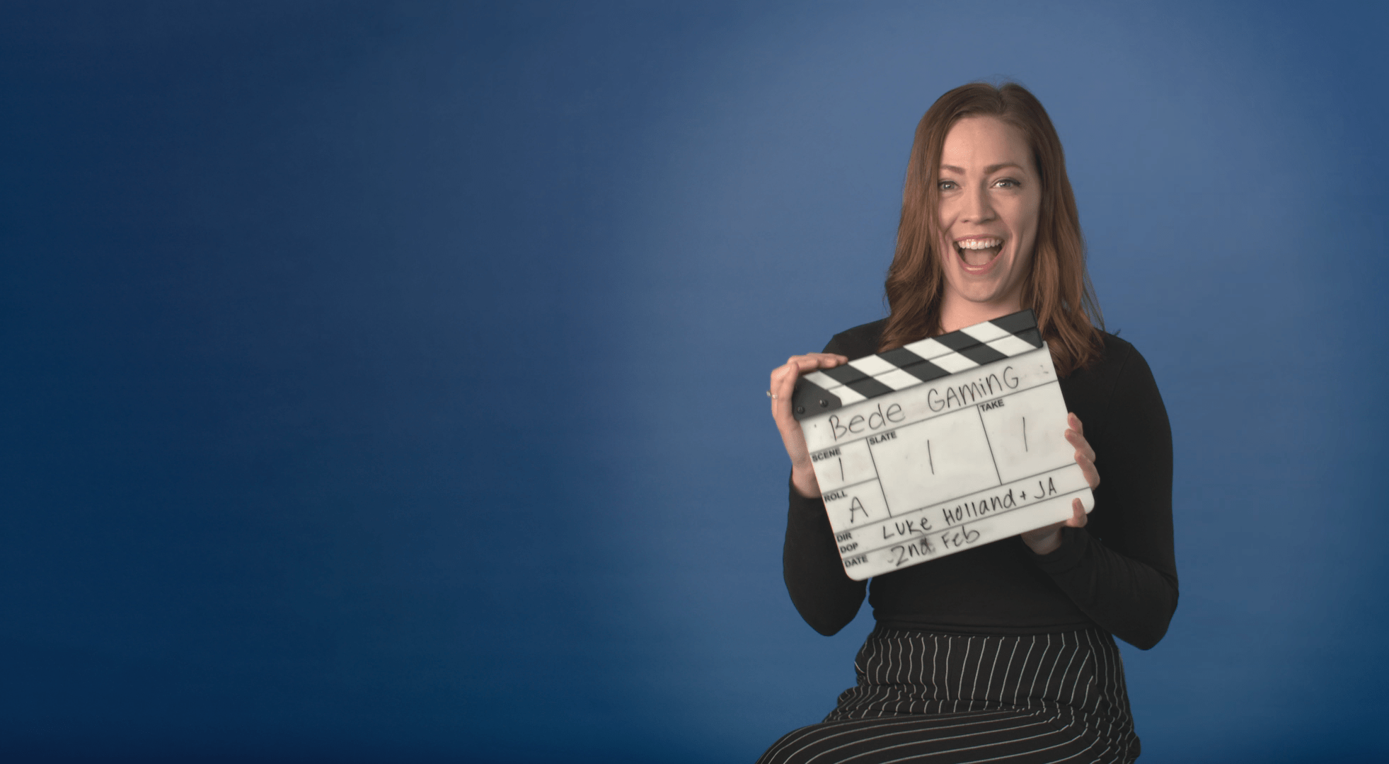 A woman poses with a Clapperboard whilst sat in front of a bluescreen backdrop.