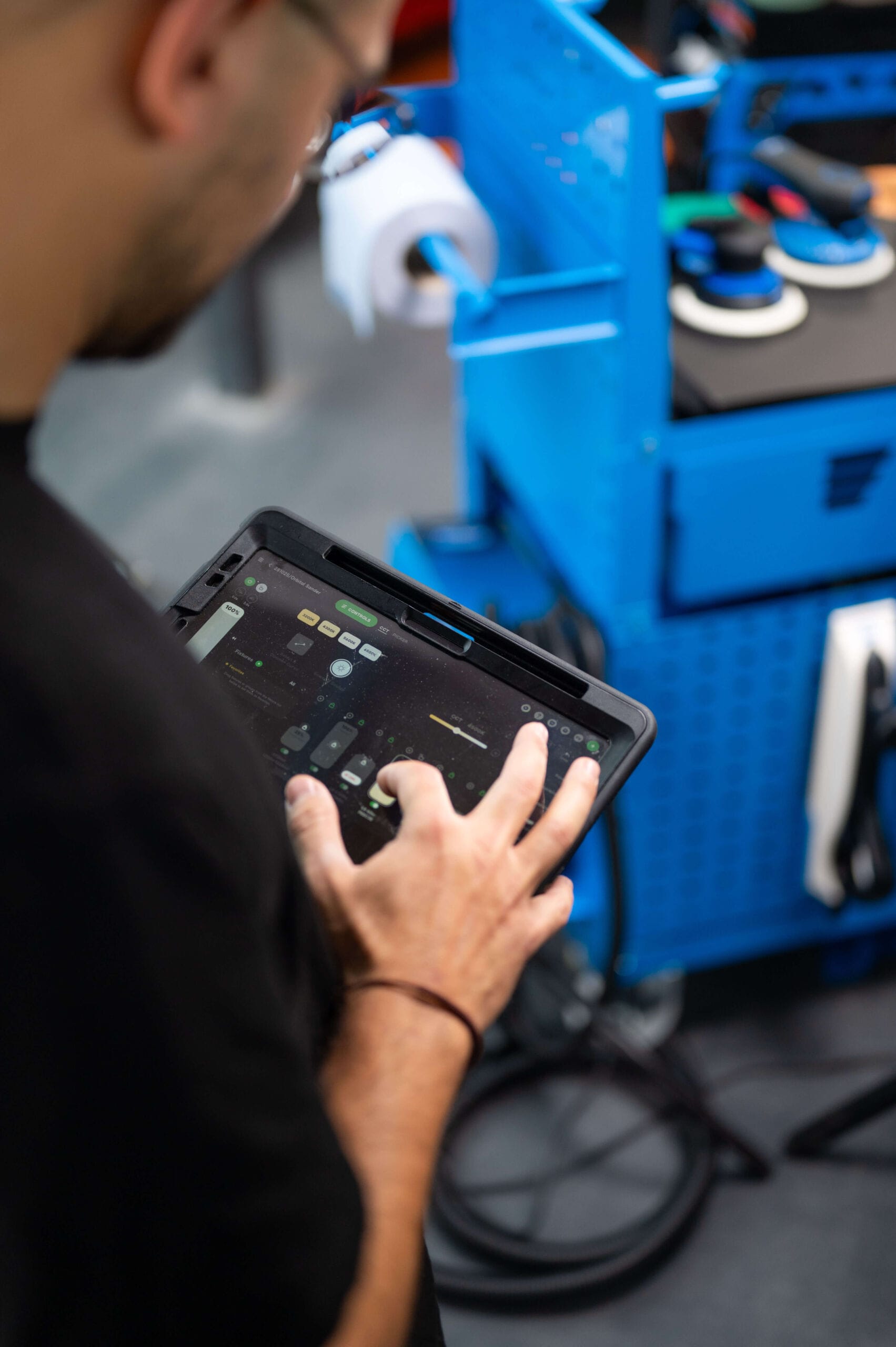 A person using a tablet to control settings near a blue workstation with tools and equipment in a workshop.