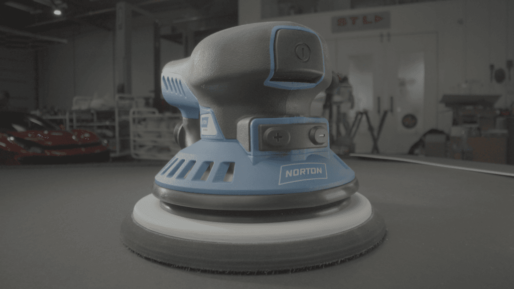 A blue and black orbital sander is positioned on a flat surface, with tools and a red car visible in the background.