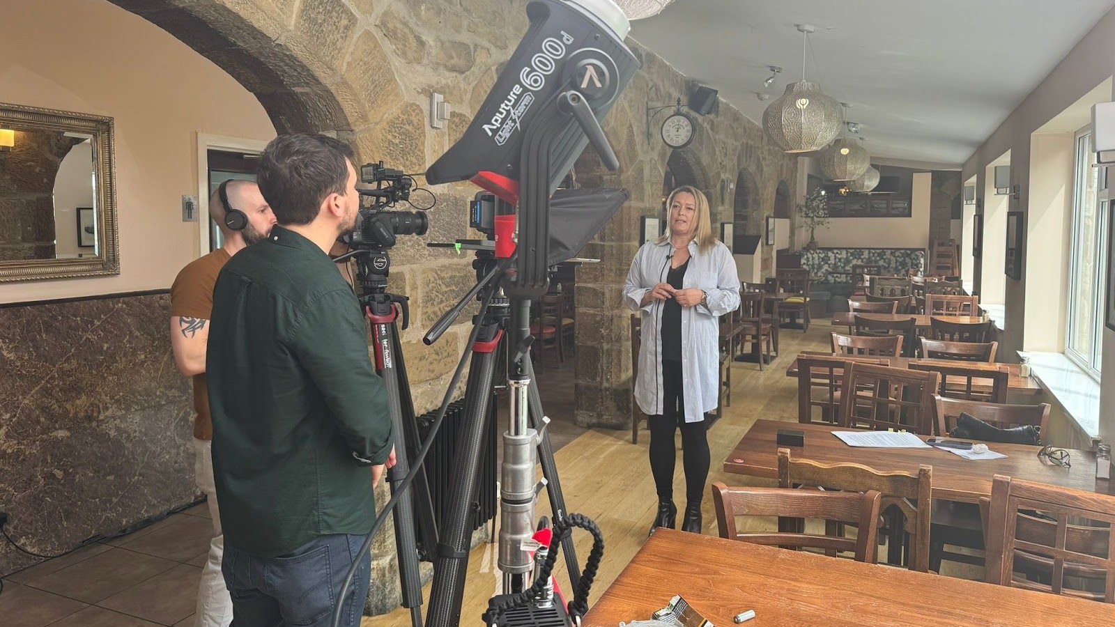 Presenter talking to camera in a lit restaurant, with a camera, a large light and crew in shot