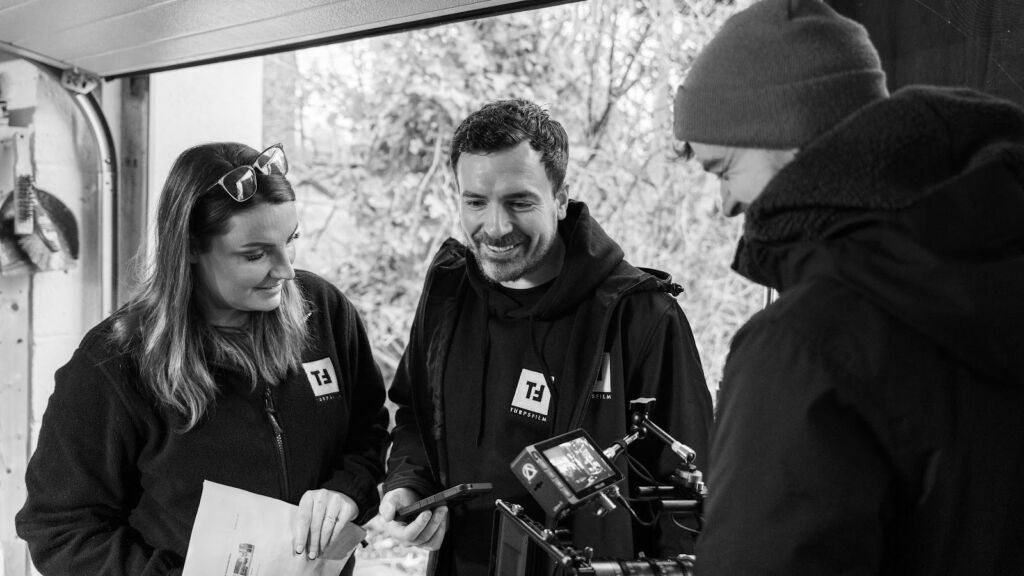 Two turps film crew members review a shot on a camera monitor, with a third crew member beside them, in black and white