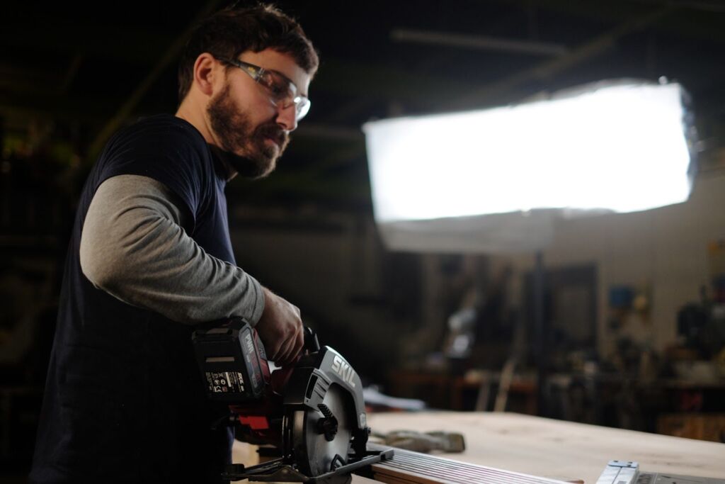 A woodworker in safety glasses demonstrating a circular saw, lit by a large softbox in a workshop