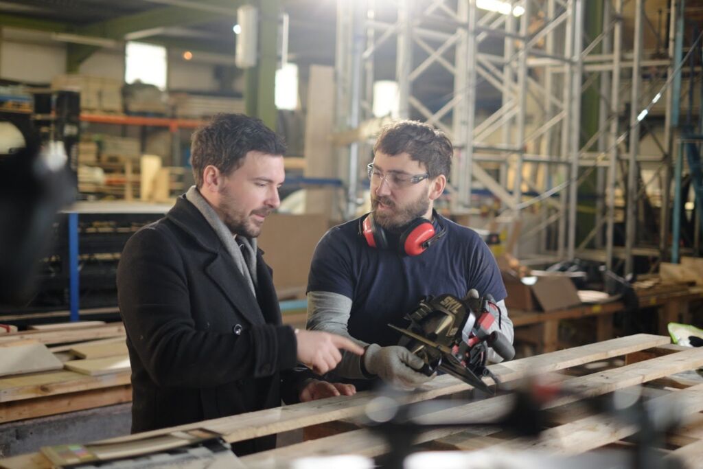 An expert in ear defenders showing a presenter how a power tool works at a workshop bench, with a camera in the foreground