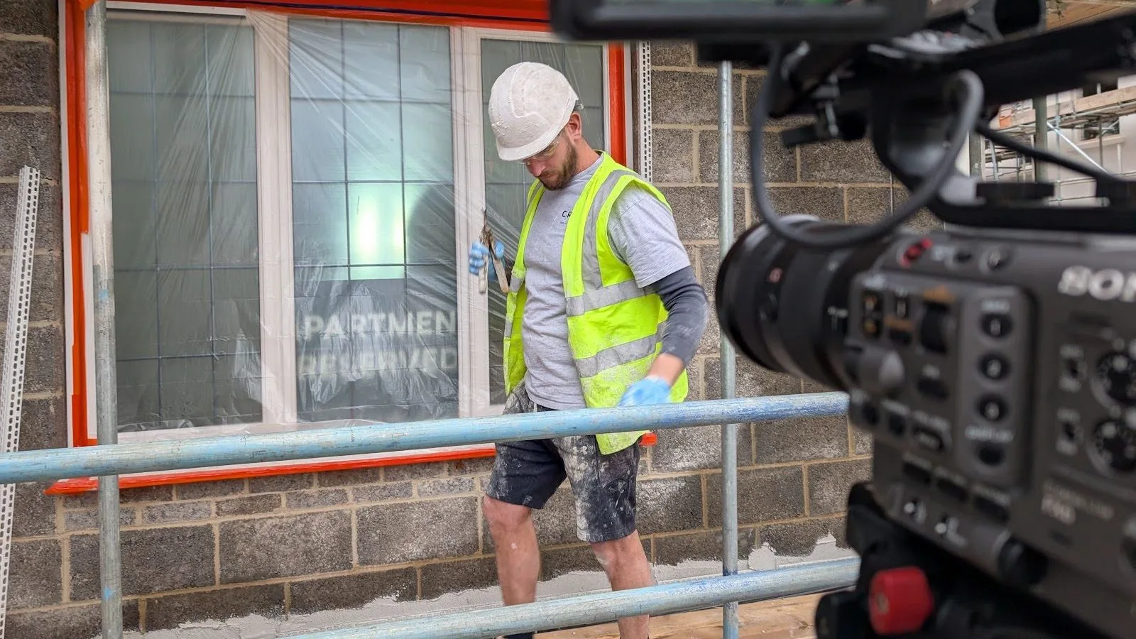 A construction worker in a hard hat and hi-vis being filmed on scaffolding, with the cinema camera in the foreground